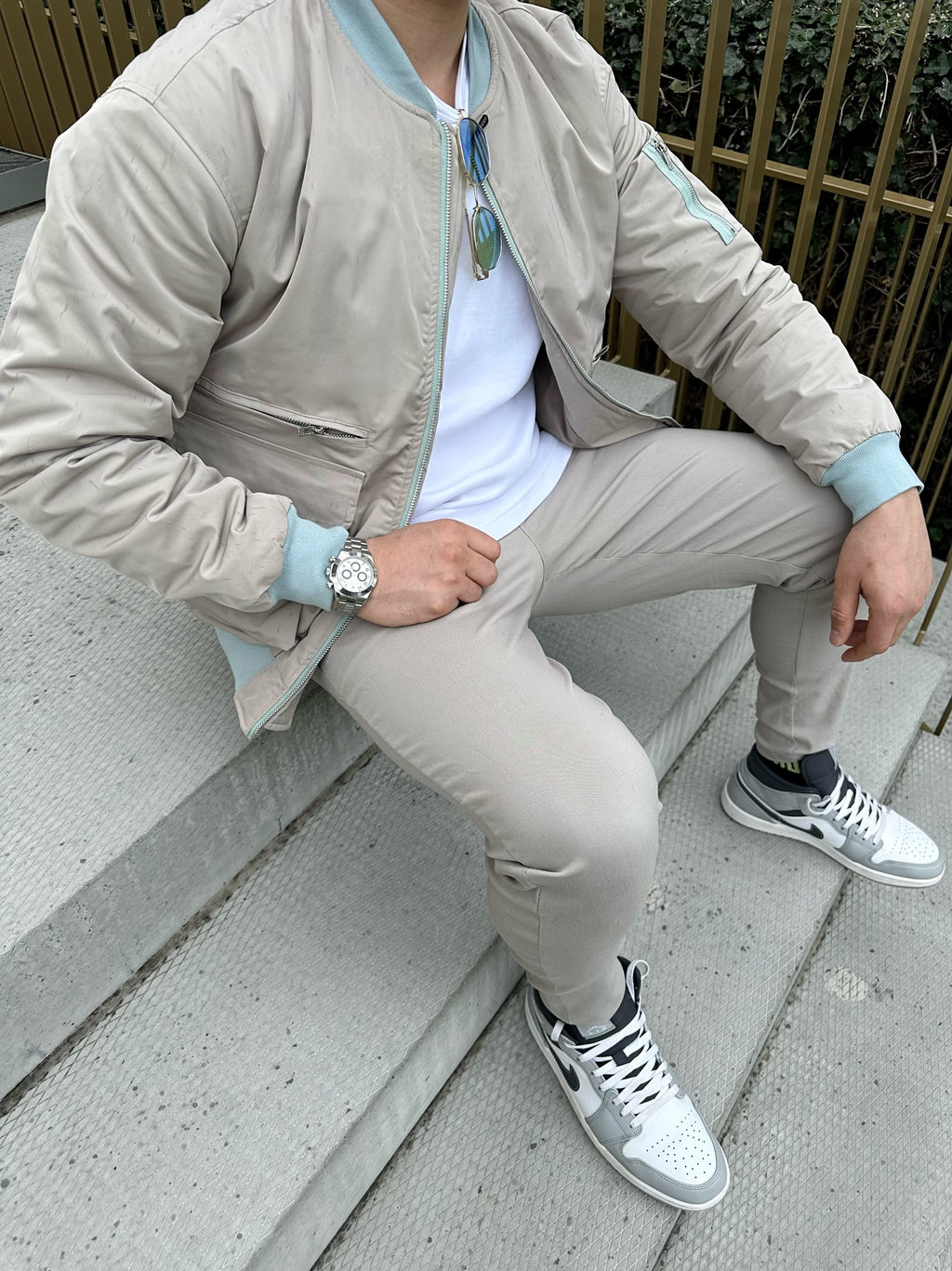 Man zit op traptrede terwijl hij een beige bomberjack draagt met witte t=shirt en beige broek, voorzijde close-up.