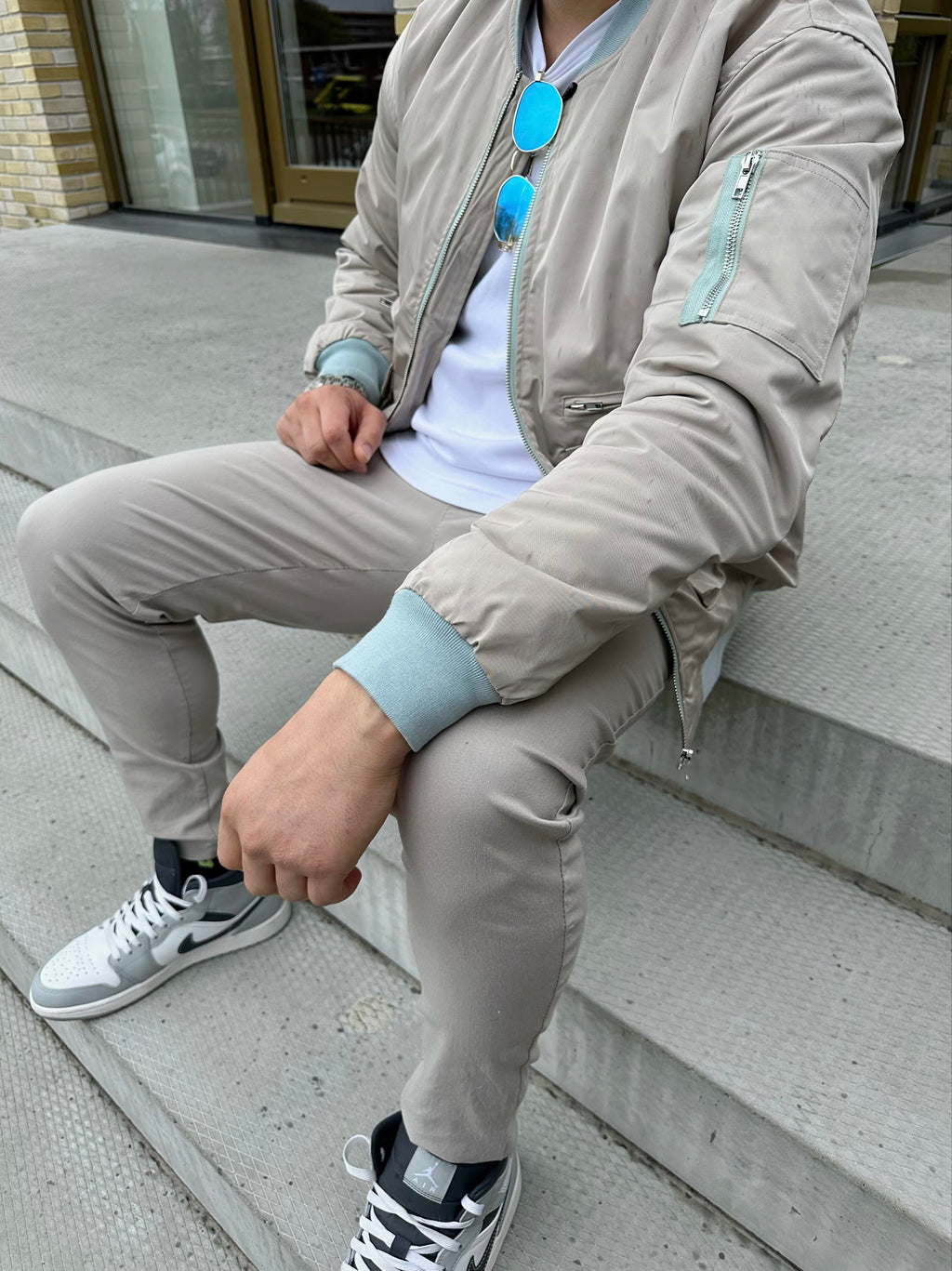Zijkant van een man die op een trap zit met beige bomberjack, witte t-shirt en beige chino.
